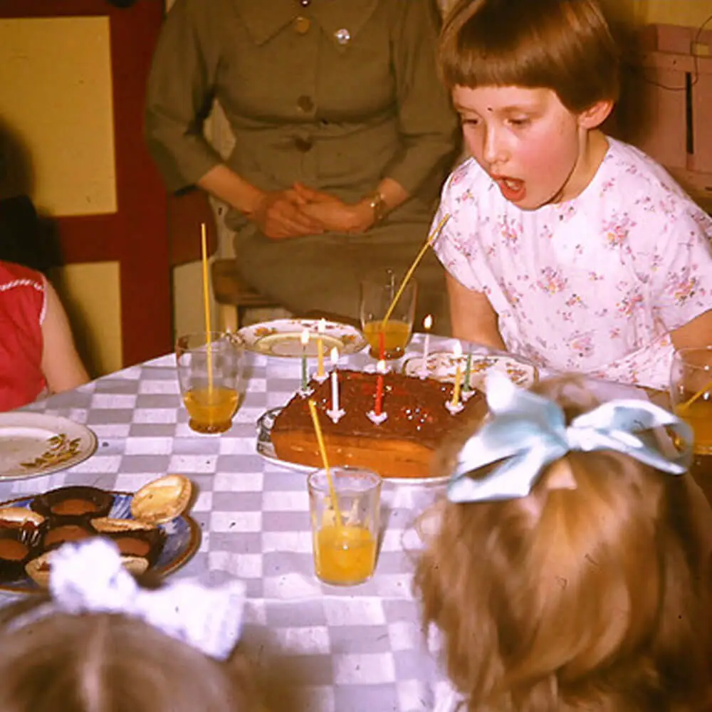 Vintage 1950s children's birthday party photo showing a girl blowing out candles on a homemade chocolate cake.