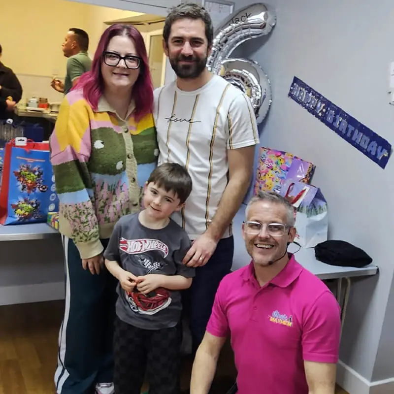 A Music and Mayhem children's entertainer in a pink polo shirt kneeling and smiling next to a young birthday boy and his parents at Preston Cricket Club. A silver number 6 balloon and Hot Wheels themed party bags are visible in the background.