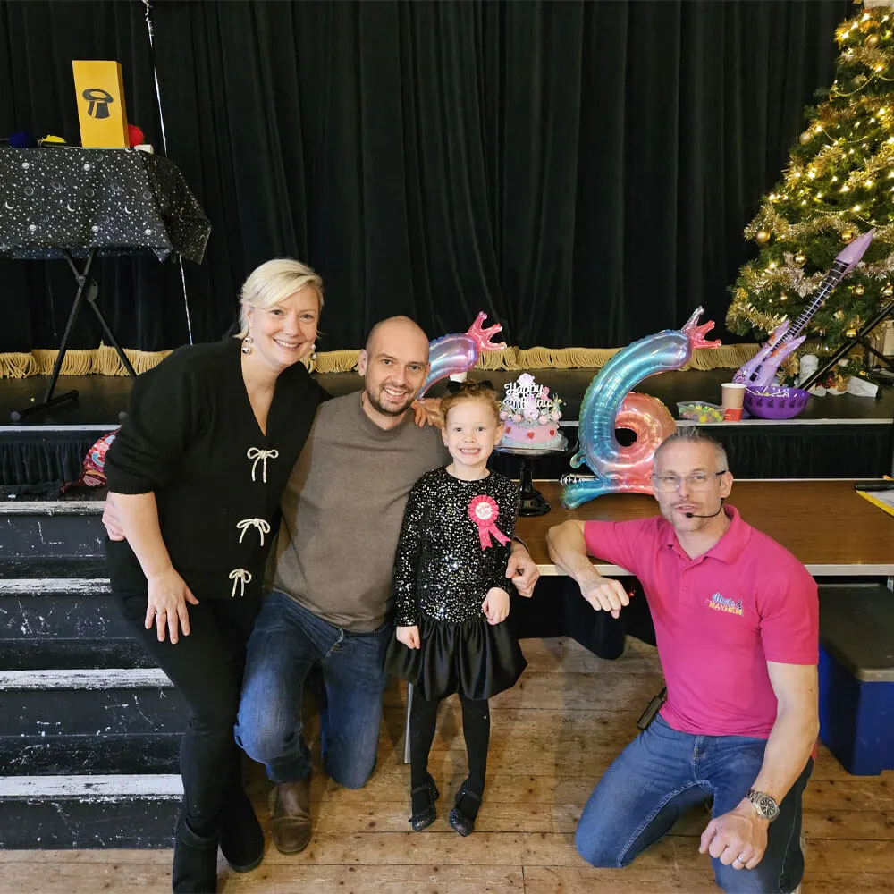 A smiling family posing with a Music and Mayhem children's entertainer in a pink branded polo shirt at a birthday party. The birthday girl stands center stage wearing a sparkly dress and badge, flanked by her parents and the entertainer, with a birthday cake, large balloon, and magic show props in the background.