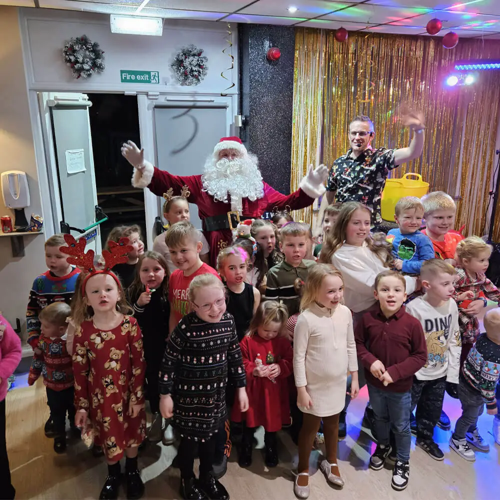 Music and Mayhem entertainer with a microphone posing with Santa Claus, children, and guests at a festive Christmas party event.