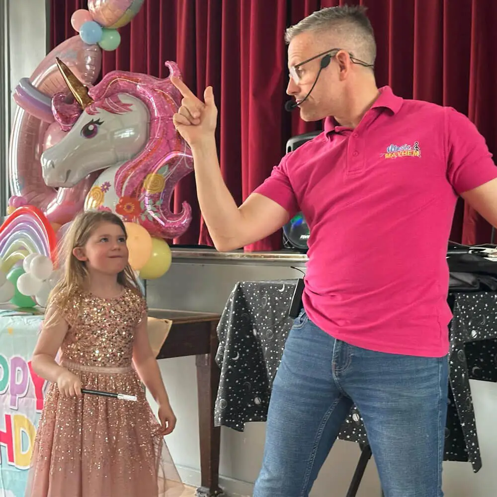 A Music and Mayhem entertainer in a pink polo shirt hosting a magic show for a child holding a wand at a festive December birthday party.