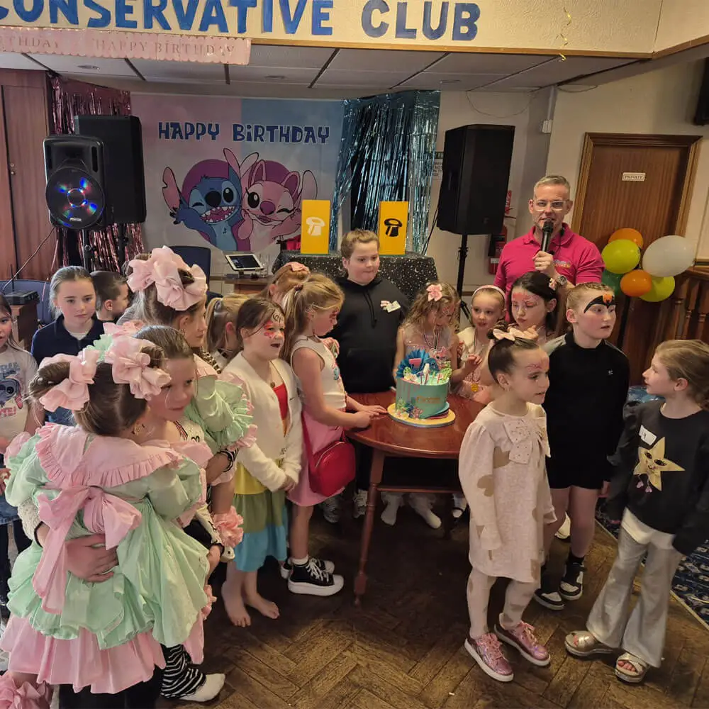 Music and Mayhem host leading a group of children singing happy birthday around a Stitch-themed cake at a party venue during the Christmas season.