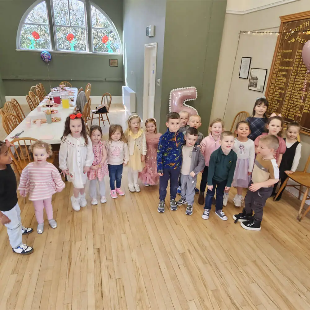 A group of children standing together for a photo near a large pink number five balloon and a decorated party food table at a village hall birthday celebration.