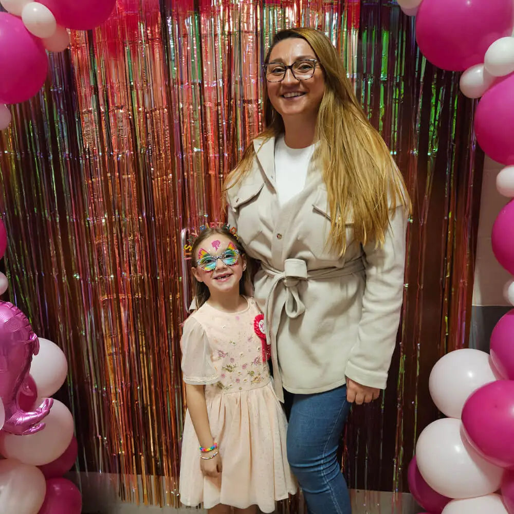 A smiling young girl with colourful butterfly face paint stands next to a woman in front of a rose gold tinsel curtain and pink and white balloon arch at a birthday party. The girl is wearing a pale pink floral dress and a birthday headband, while the woman wears glasses and a beige belted jacket.