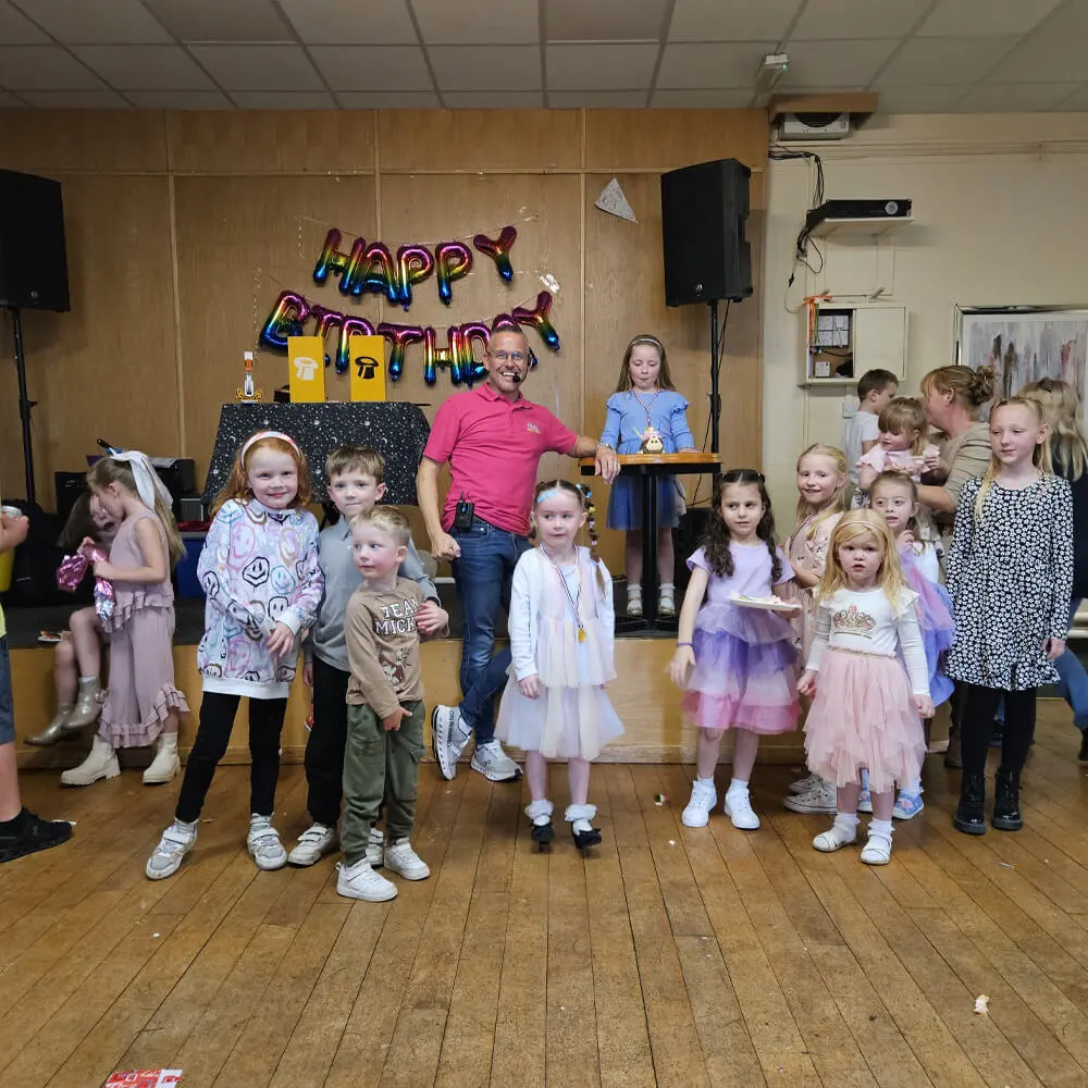 Music and Mayhem children's entertainer standing with a group of kids on the dance floor in front of a happy birthday balloon banner.