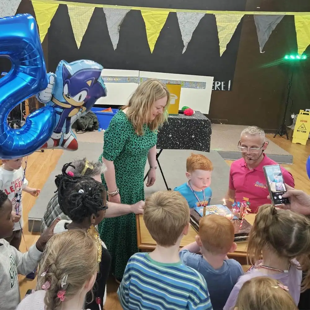 Children's party entertainer from Music & Mayhem hosting a 5th birthday party in Lancashire as a young boy blows out his birthday cake candles surrounded by friends.