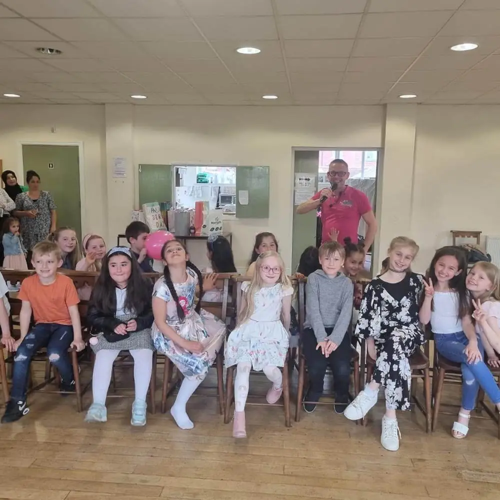 Children's party entertainer from Music & Mayhem hosting a 7th birthday party celebration with a group of children seated on chairs in a community hall in Lancashire.